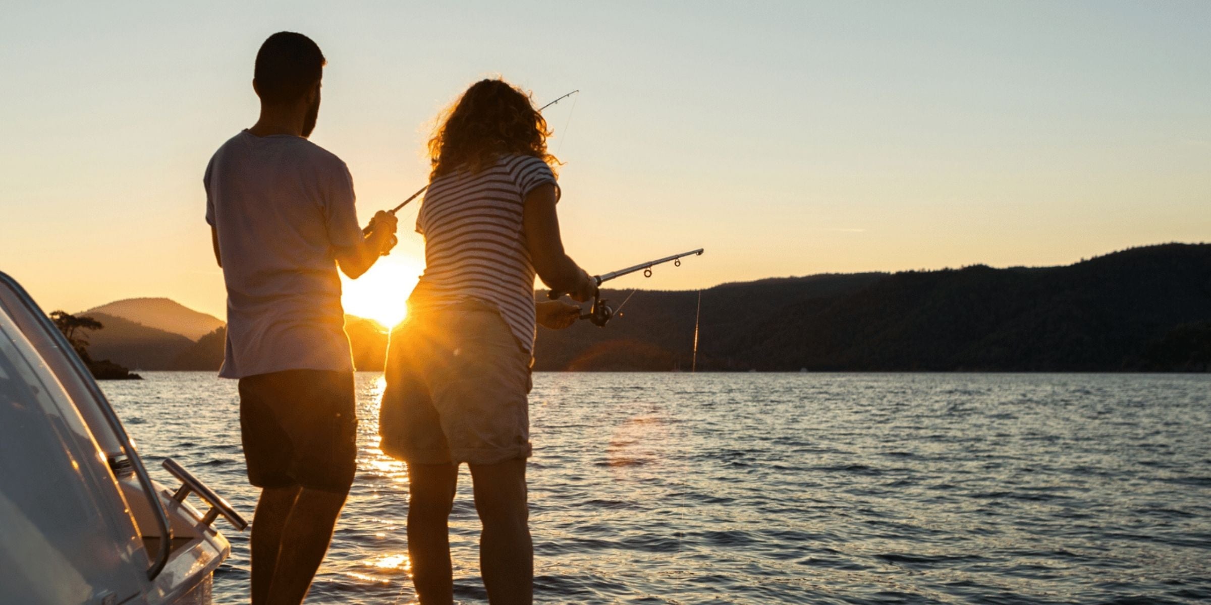 a serene scene during sunset. Two people are fishing off the side of a boat, their silhouettes cast against the golden sun