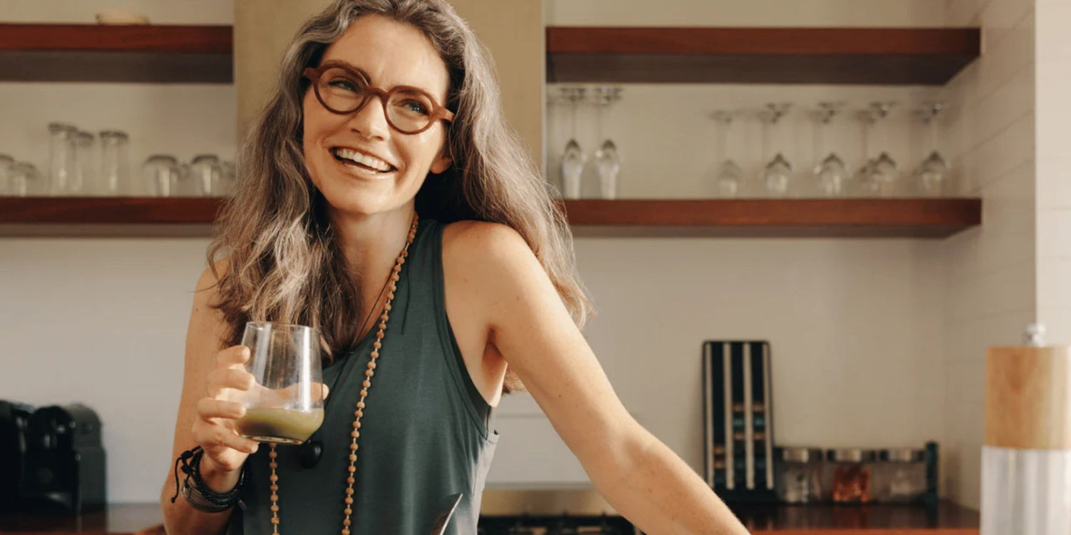 a mature woman standing in her kitchen drinking a green smoothie